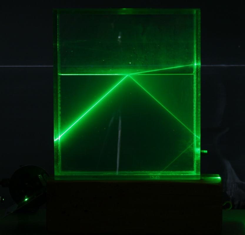 Green laser beam approaching the water surface at a larger angle of incidence, with bright reflected beam forming a V-pattern in the water and a refracted beam exiting to the upper right