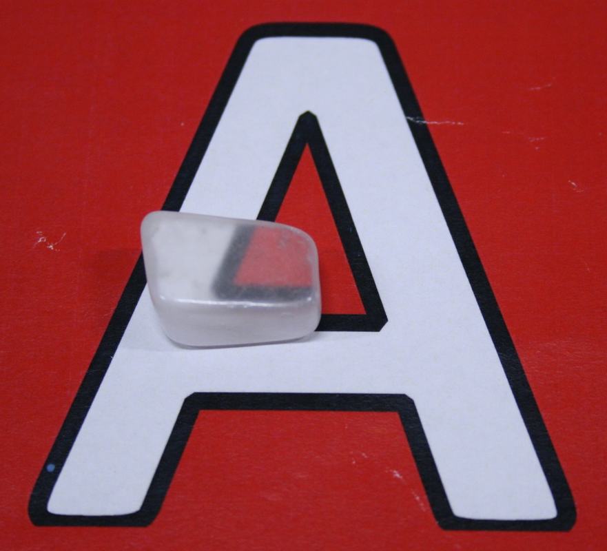 Small translucent ulexite specimen (~1x1x2 cm) resting on a large white letter A on a red background, with part of the letter's edge visible through the top of the stone