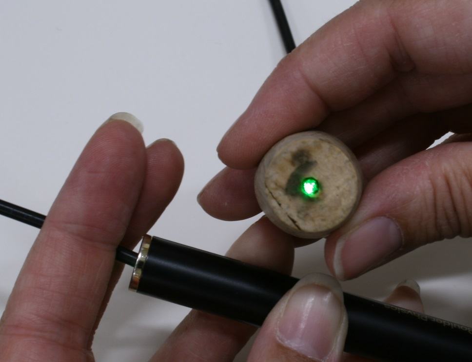 Hand holding a round fiber bundle end showing a bright green light spot emerging, with the other hand holding the black cable connector