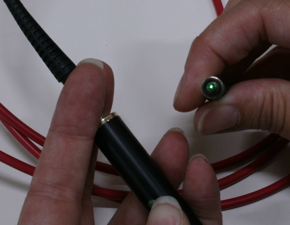 Hands holding black fiber optic cable with green light emerging from the polished connector end, red cables visible in background