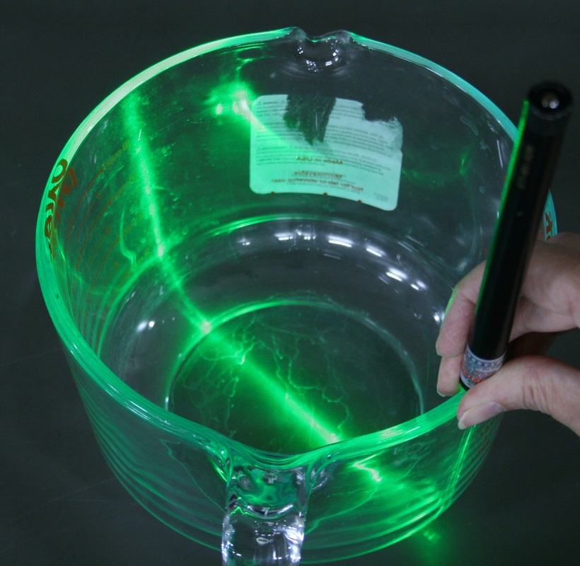 Hand holding a green laser pointer aimed into the rim of a glass measuring cup, green light visible propagating through the glass walls