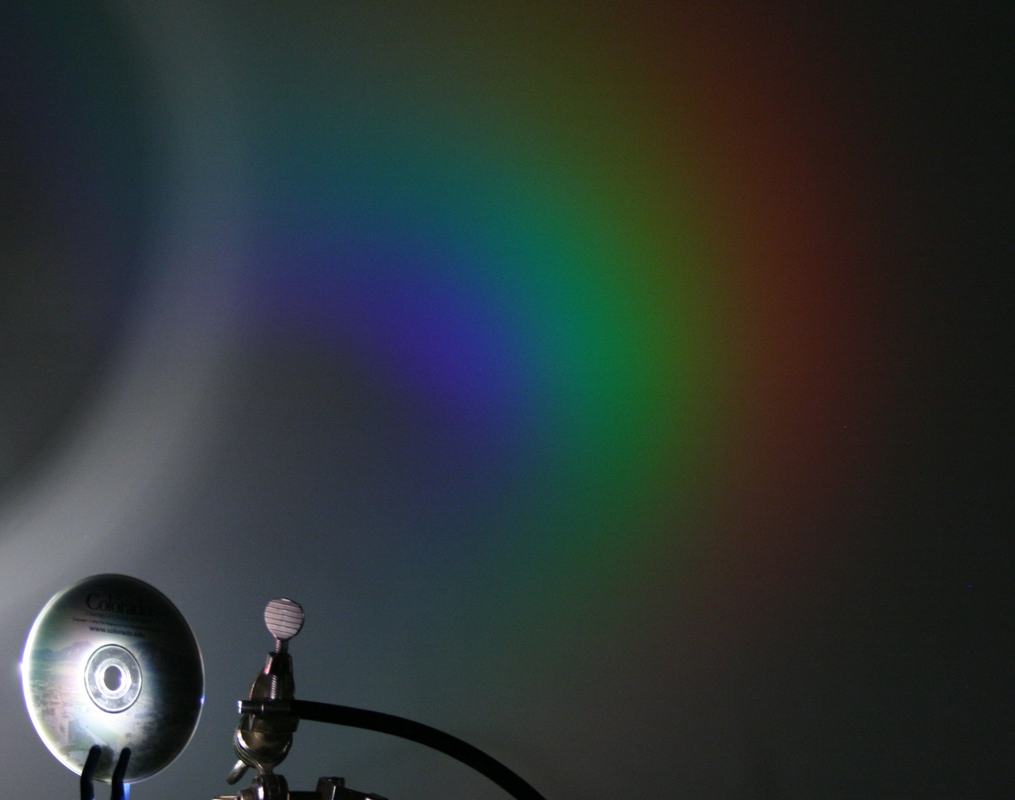 Closer view of the CD and light source, showing a vivid diffracted spectrum with violet, blue, green, and red bands on the wall