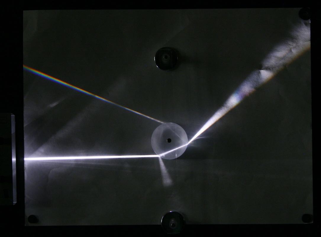 Small circular acrylic disk on blackboard optics board with white light beam entering from the left, producing a dispersed rainbow spectrum to the upper left and a refracted beam to the upper right