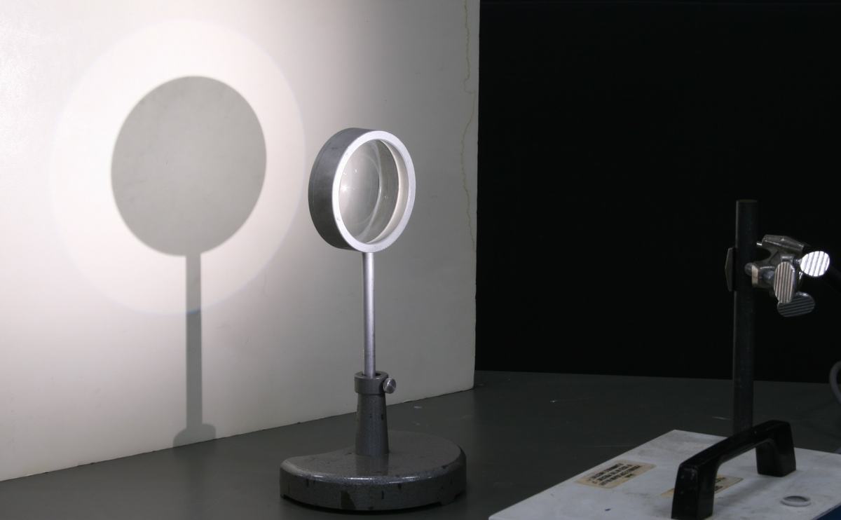 Concave lens mounted on a post stand casting a circular shadow on a white screen, with a bright light source visible at the right edge