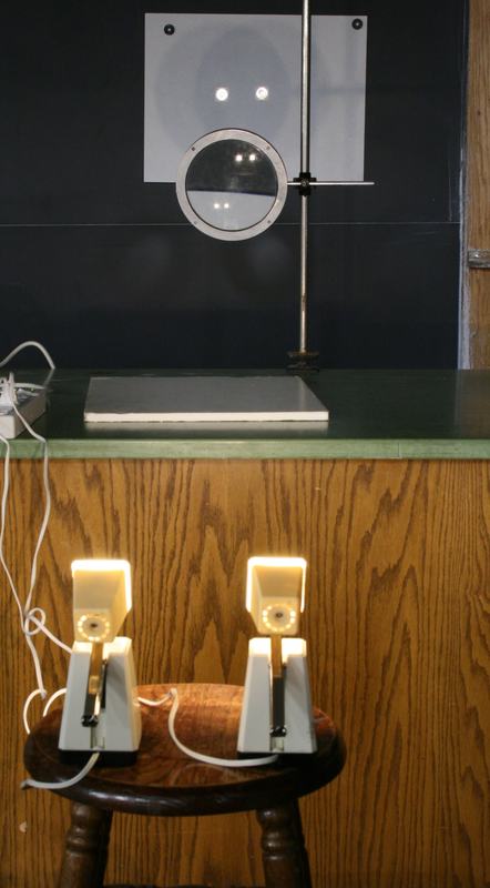 Large convex lens clamped to a vertical rod on the lecture bench with a gray mounting plate, two illuminated desk lamps on a wooden stool positioned below
