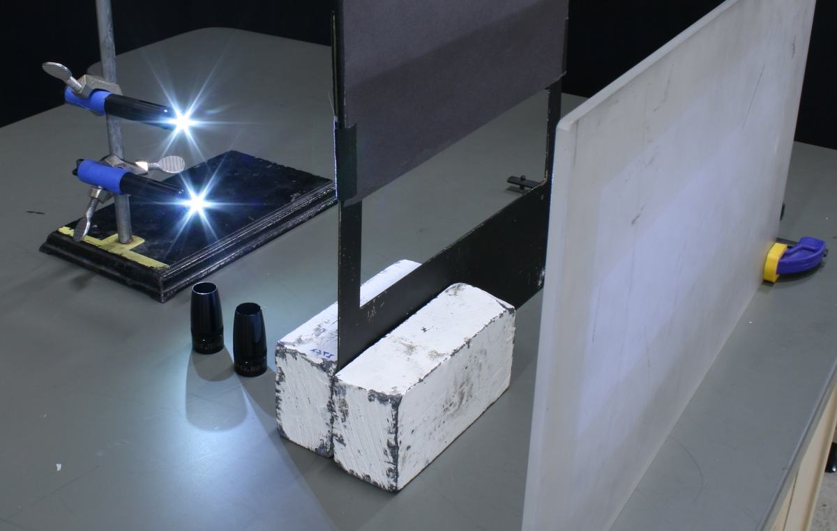 Two flashlights clamped to a ring stand at left, dark rectangular mask propped upright with a white-painted lead brick, translucent screen at right, flashlight caps on bench