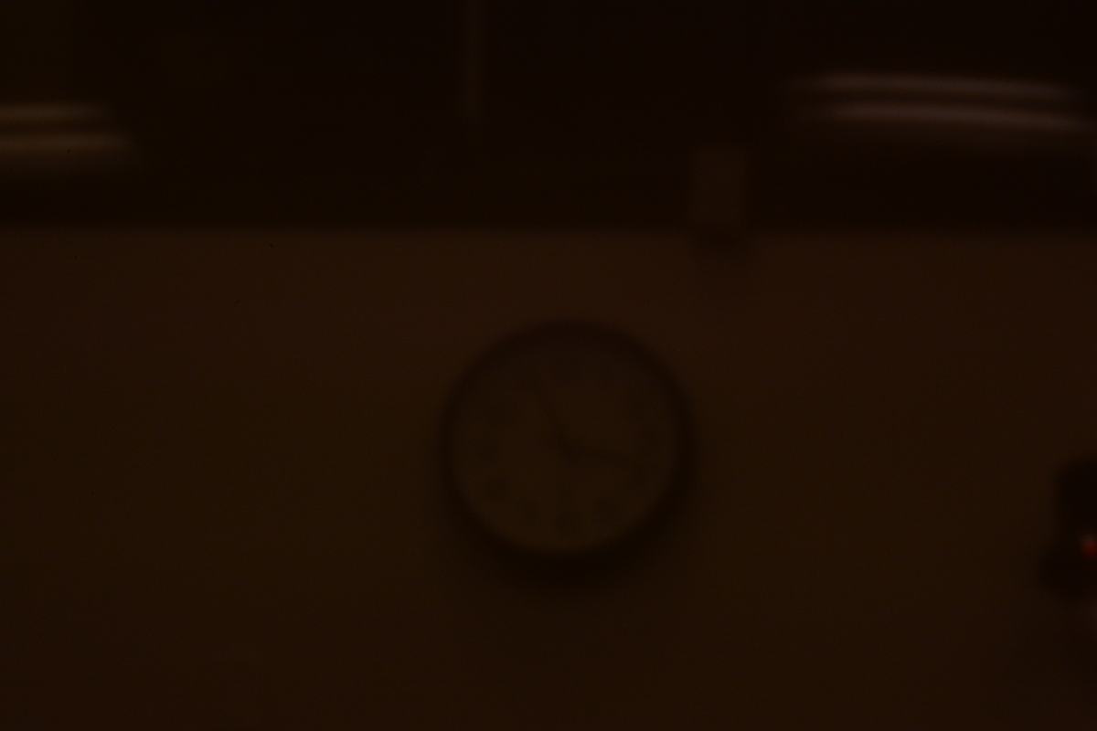 Very dark photograph of a lecture hall wall clock through the smallest pinhole; clock outline is sharper but image is severely underexposed