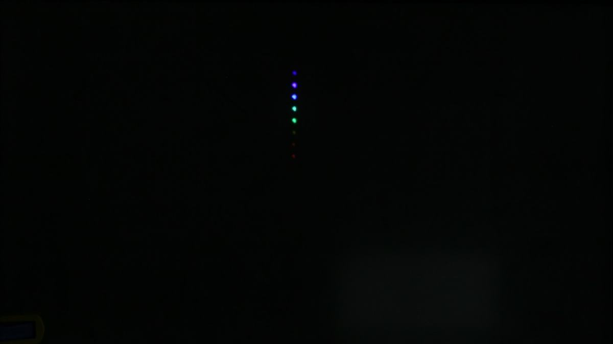 Vertical column of colored dots on dark screen — violet, blue, green, yellow, red from top to bottom — projected from LED tower through pinhole