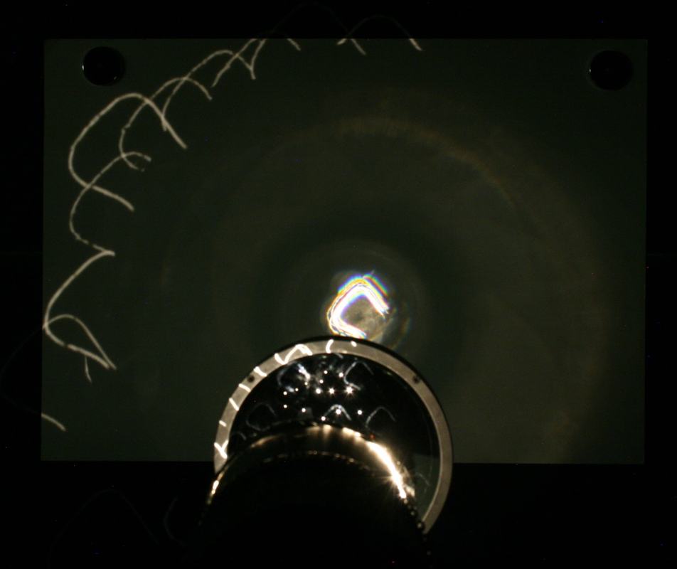 Large lens focuses all pinhole images to a single bright point with chromatic aberration color fringes; a few filament images remain visible at upper left