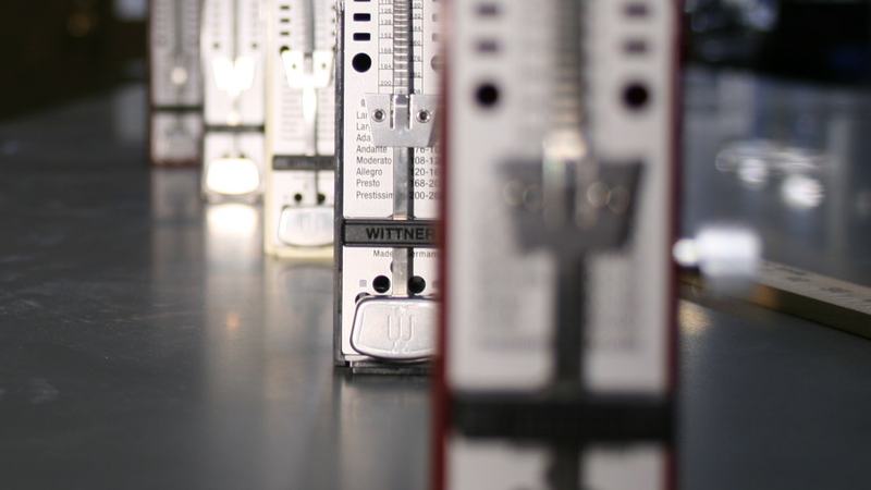 Row of Wittner mechanical metronomes on a lab bench shot from low angle; the nearest red-and-white metronome is sharp while background metronomes progressively blur