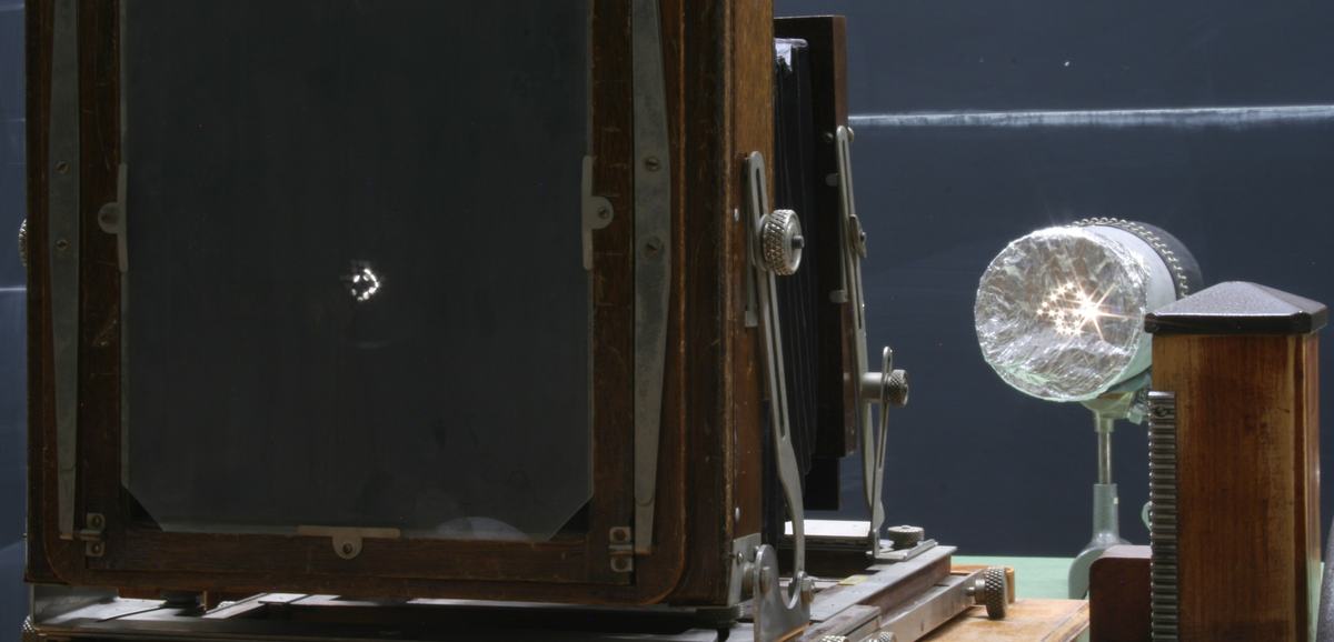 Large-format wooden view camera with ground glass plate on an optical bench, with a lamp covered in aluminum foil (pinholes visible) to the right