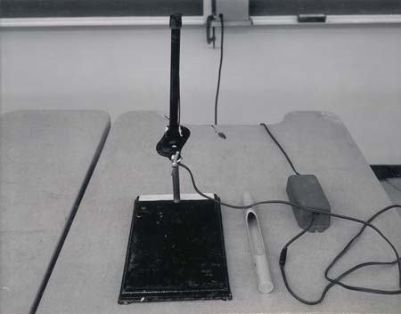 UV fluorescent tube mounted vertically on a ring stand with parallel clamp. The aluminum case and power supply sit on the table beside it.