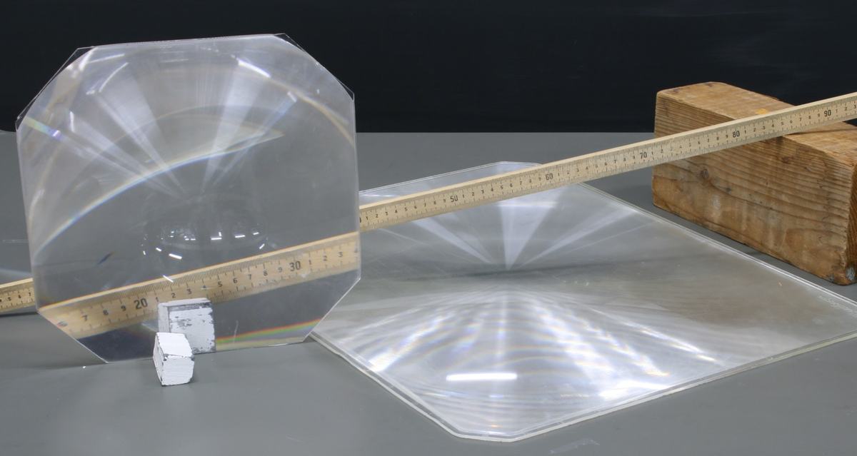 Large octagonal Fresnel lens propped upright and smaller square Fresnel lens lying flat, with a wooden ruler and small white cube for scale