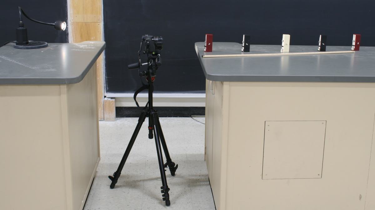 Full setup in lecture hall: camera on tripod between two tables, five metronomes spaced along a meter stick on the far table, desk lamp on the left table providing illumination