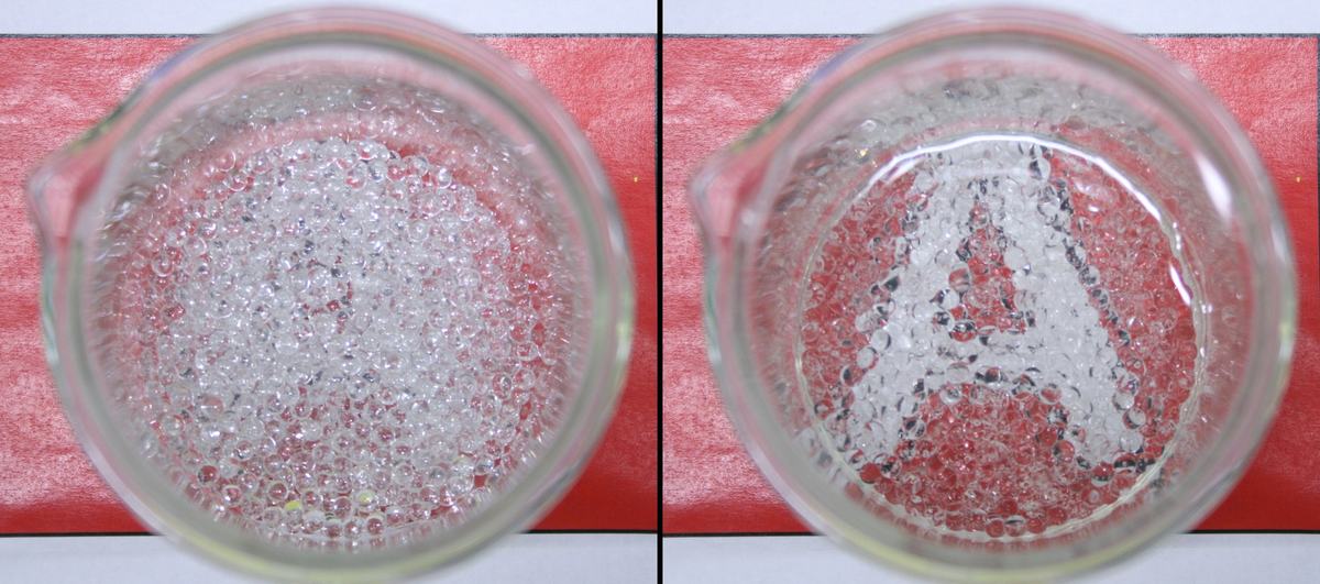 Top-down view of two beakers on red paper with a letter printed on it. Left: dry glass spheres obscure the letter. Right: glass spheres submerged in water become transparent, revealing the letter beneath