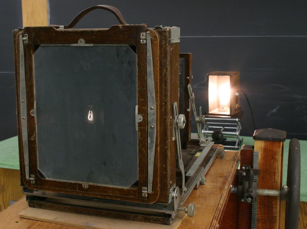Rear view of view camera showing ground glass screen with inverted filament image visible, carbon filament lamp glowing behind the camera on the rail