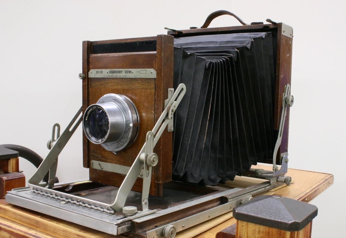 Wooden 8x10 view camera with black bellows extended, metal lens on front standard, mounted on wooden base with metal rails
