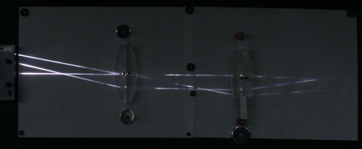Two convex lenses on a magnetic whiteboard with diverging rays from a projector at left converging between the lenses and then diverging again through the second lens