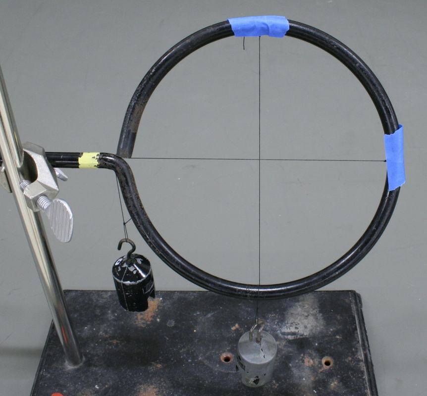 Close-up of black ring with blue tape holding threads, perpendicular threads crossing at center, mounted on ring stand clamp with hanging weight for tension