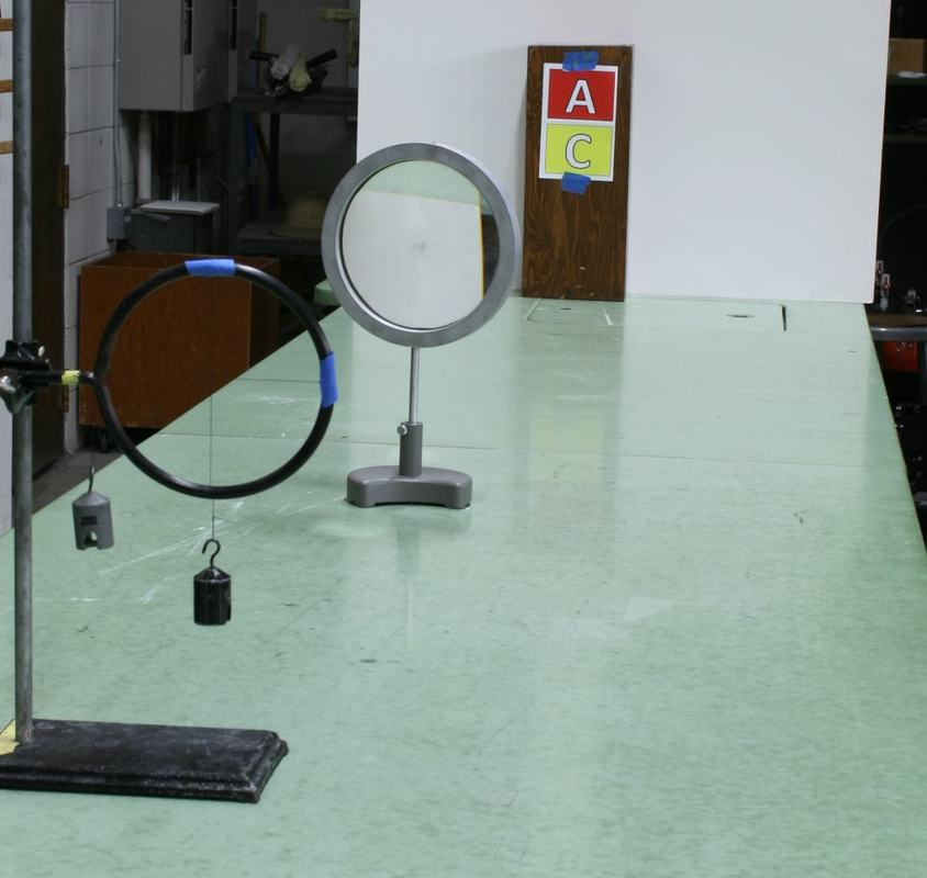 Wide view of setup on green bench: crosshair ring on ring stand at left, convex lens on tabletop mount in center, wooden block with colorful letters A and C at right end of bench