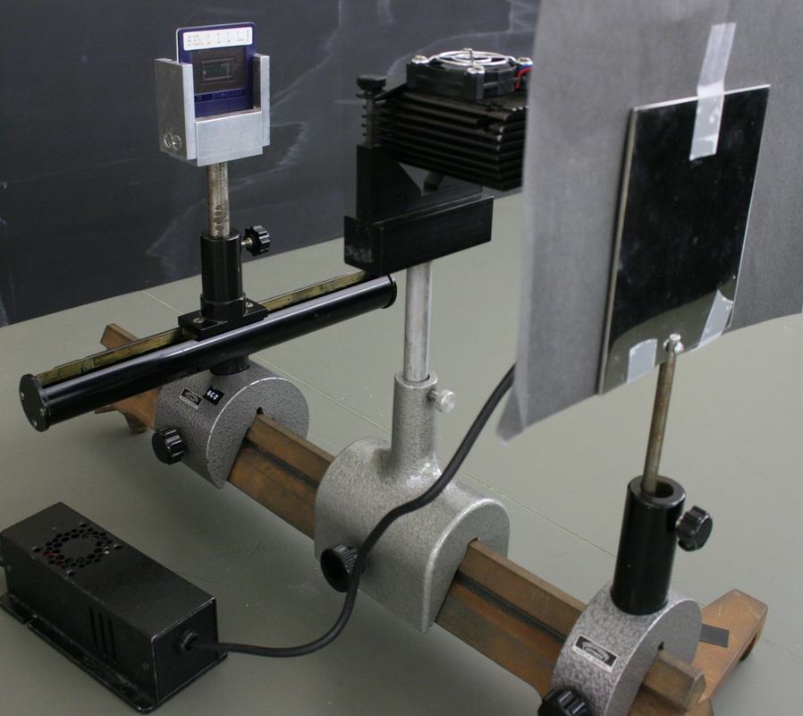 Optical rail setup with slit slide in adjustable carriage on the left, laser with heatsink in the center, and construction-paper reflection shield on polished steel plate at the right end, with a laser power supply on the table