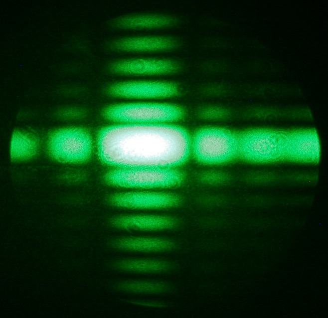 Green laser diffraction pattern with wider horizontal spread and fewer vertical fringes, showing an asymmetric cross pattern