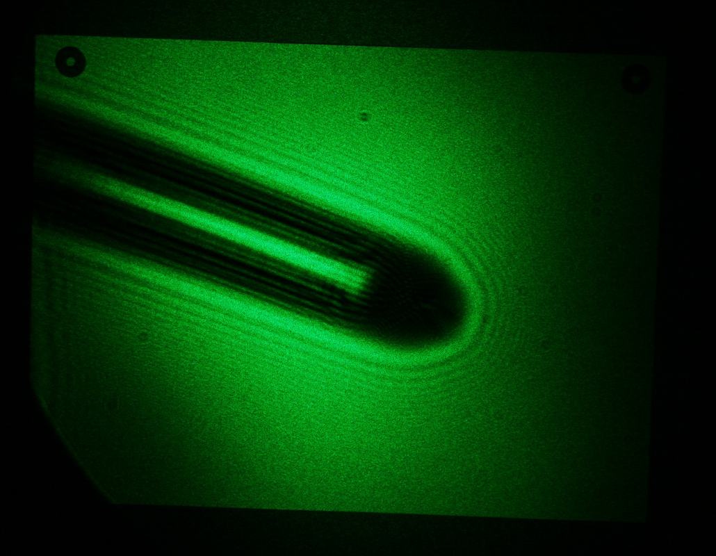 Green laser diffraction pattern from a needle showing elongated dark shadow surrounded by curved Fresnel fringes