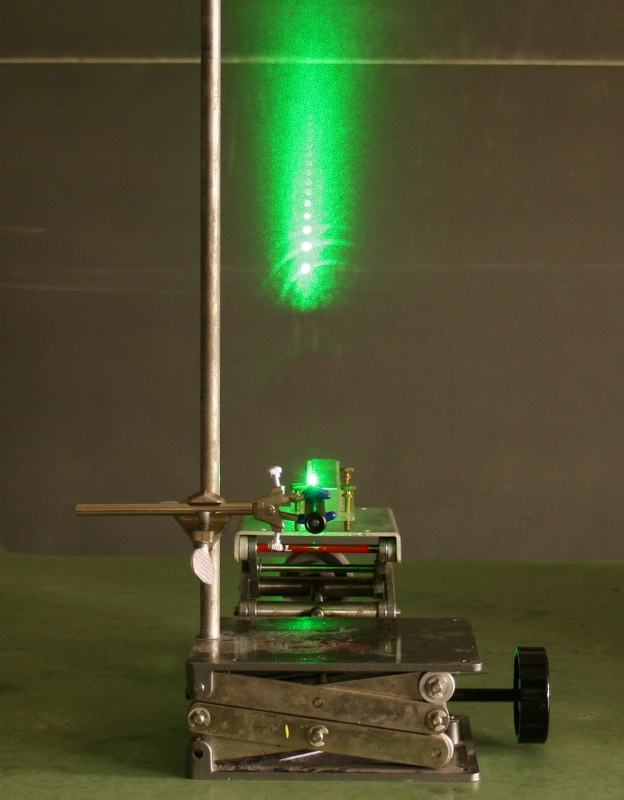 Green laser on a lab jack with beam grazing a machinist's scale, producing a vertical column of bright diffraction spots on the wall above; a ring stand rod is visible behind the setup