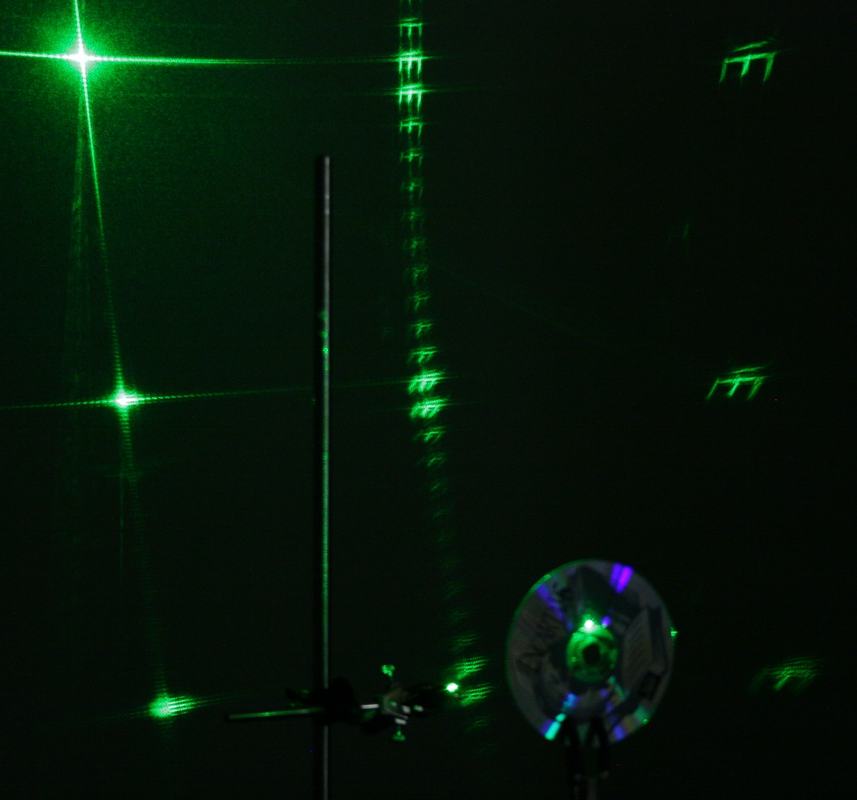 Green laser diffraction from CD showing cross-shaped pattern with multiple orders on wall, and imaged letters from manufacturer text visible at upper right