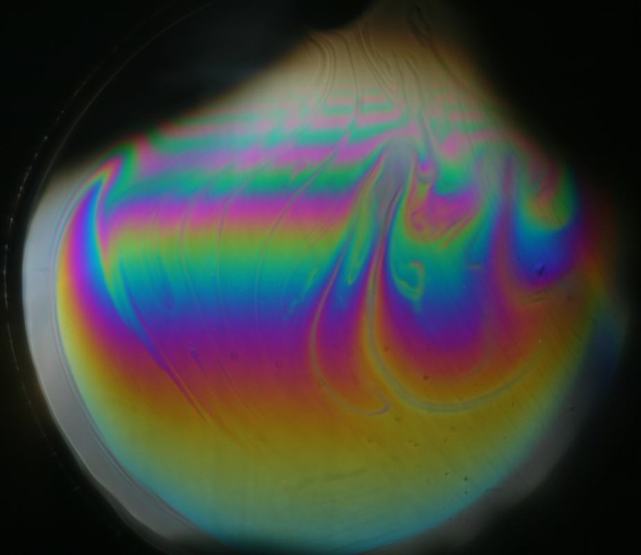 Projected soap film showing colorful interference bands of magenta, green, cyan, yellow, and blue swirling across the film against a dark background