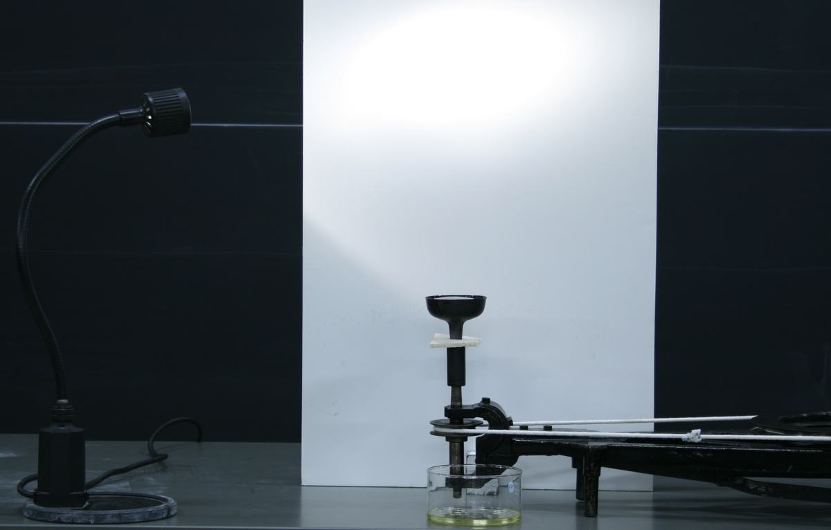 Apparatus setup: black cup on rotator arm with glass dish of soap solution below, white foam core backdrop, gooseneck lamp at left, rotator handle extending to right