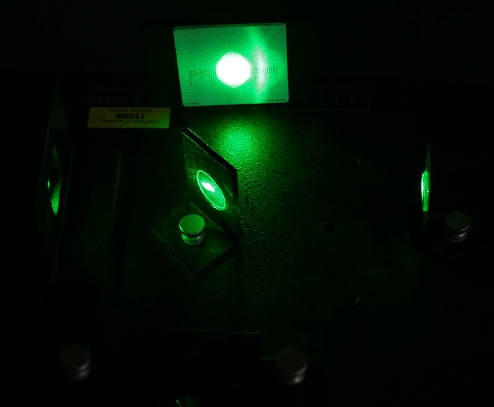 Close-up of Pasco interferometer in dark room showing beam splitter illuminated by green laser, with viewing screen above displaying bright green spot; PASCO label visible