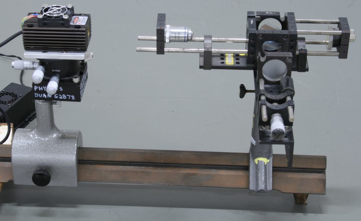 Pasco Precision Interferometer with room lights on, showing green laser with heatsink and cooling fan on left, interferometer body with micrometer and adjustment knobs on optical rail