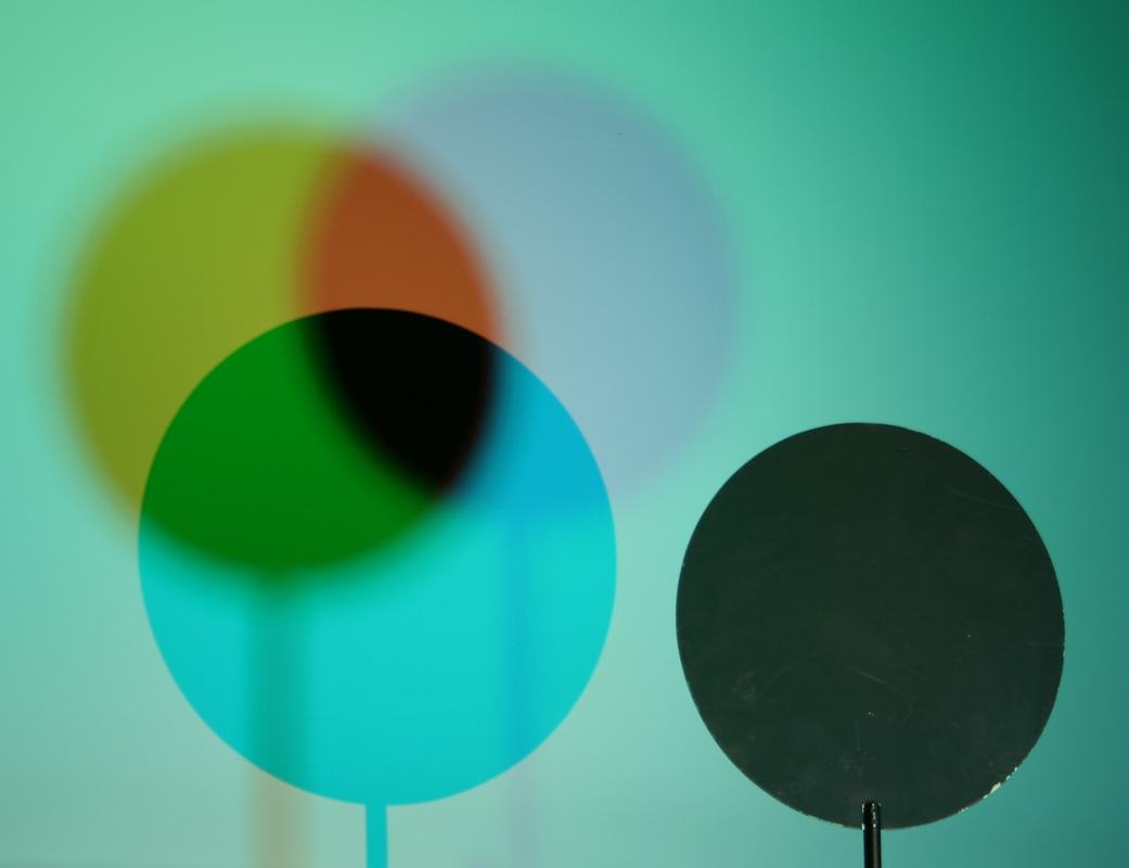 Three overlapping circular shadows on a cyan background, showing yellow, red-orange, blue-lavender, green, cyan, and black regions, with a circular barrier on a stand at right