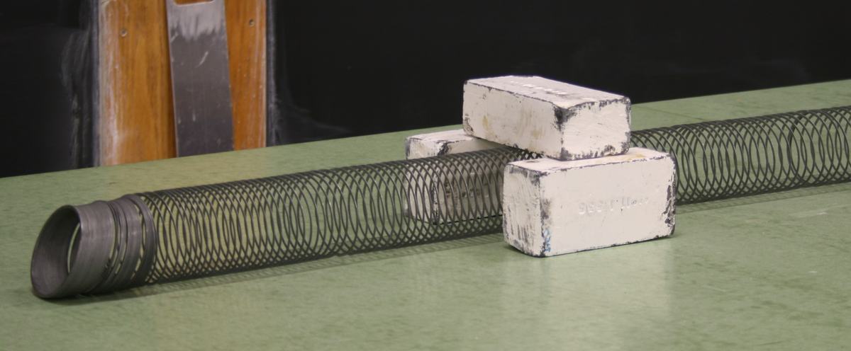 Giant metal Slinky stretched along a green demo bench with three lead bricks forming a tunnel barrier — two bricks side by side with a third across the top