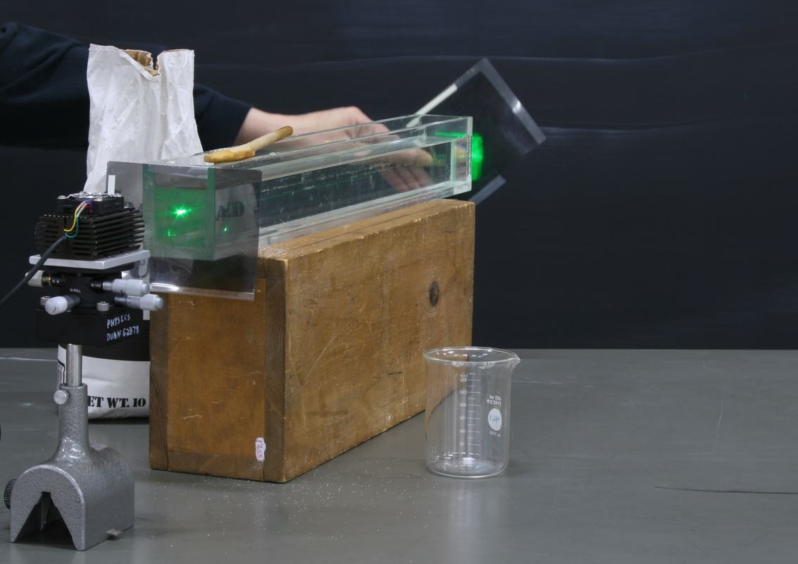 Hand holding a square polarizing filter at the exit end of the glass tank, green laser beam visible in the cleared sugar solution