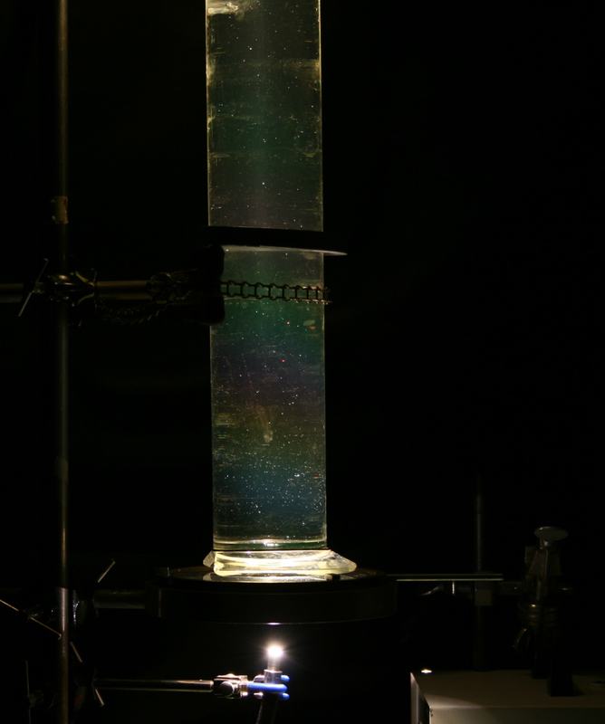 Tall glass cylinder clamped with a chain on a ring stand, illuminated from below in a darkened room, with greenish scattered light and suspended particles visible in the solution