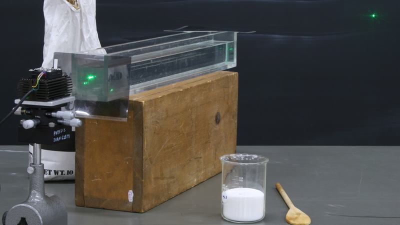 Rectangular glass tank on a large wooden block with a green laser and heat sink on the left, polarizer taped to tank entrance, beaker of sugar and wooden spoon on right