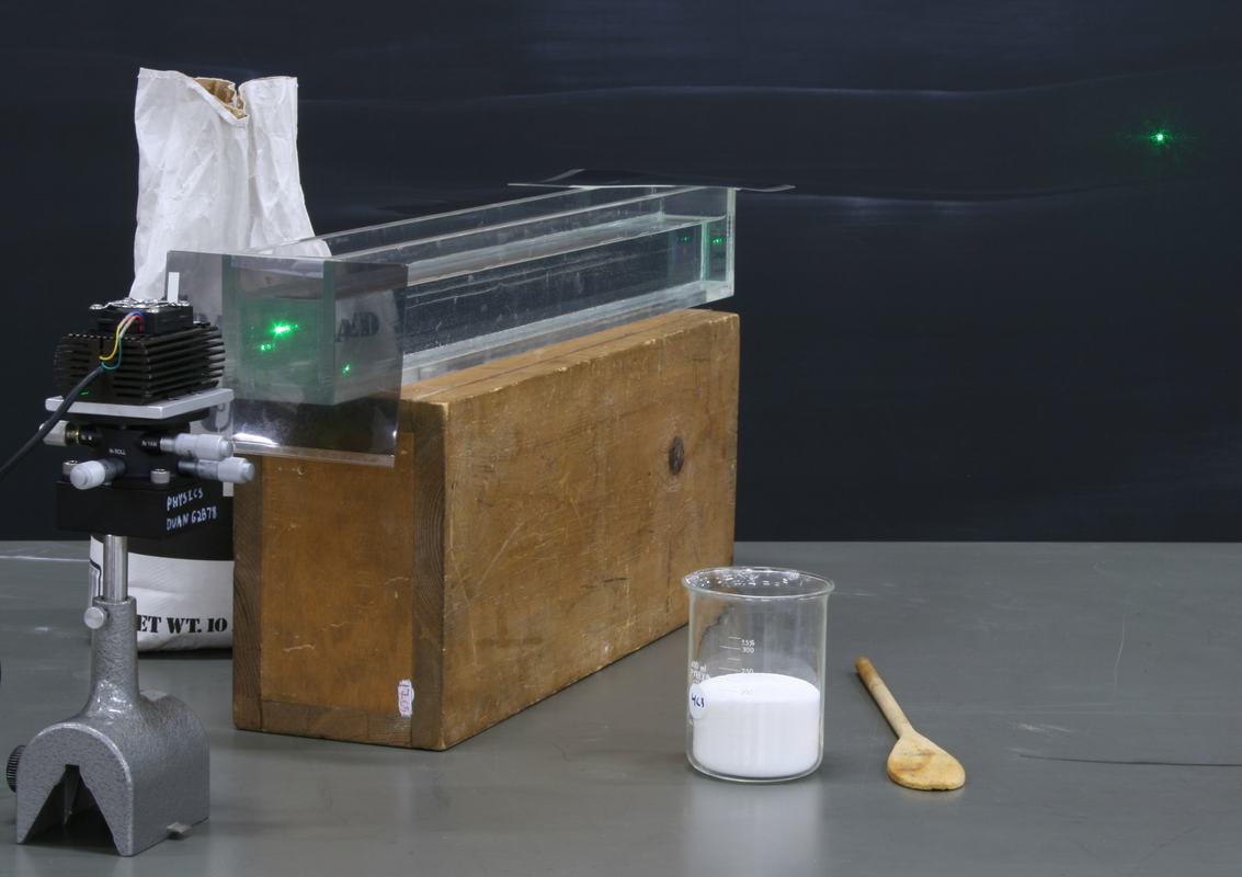 Rectangular glass tank on a large wooden block with a green laser and heat sink on the left, polarizer taped to tank entrance, beaker of sugar and wooden spoon on right