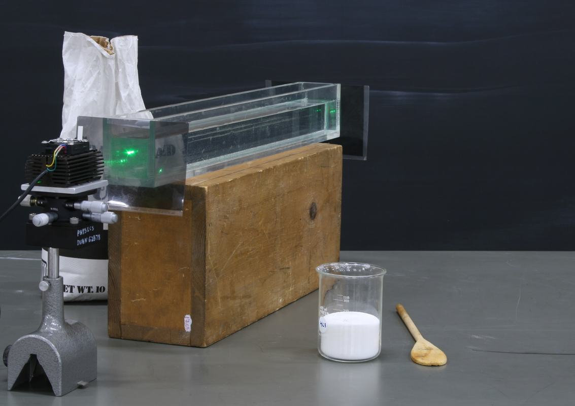 Same setup from slightly different angle showing glass tank on wood block, green laser, polarizers, beaker of sugar, and wooden spoon