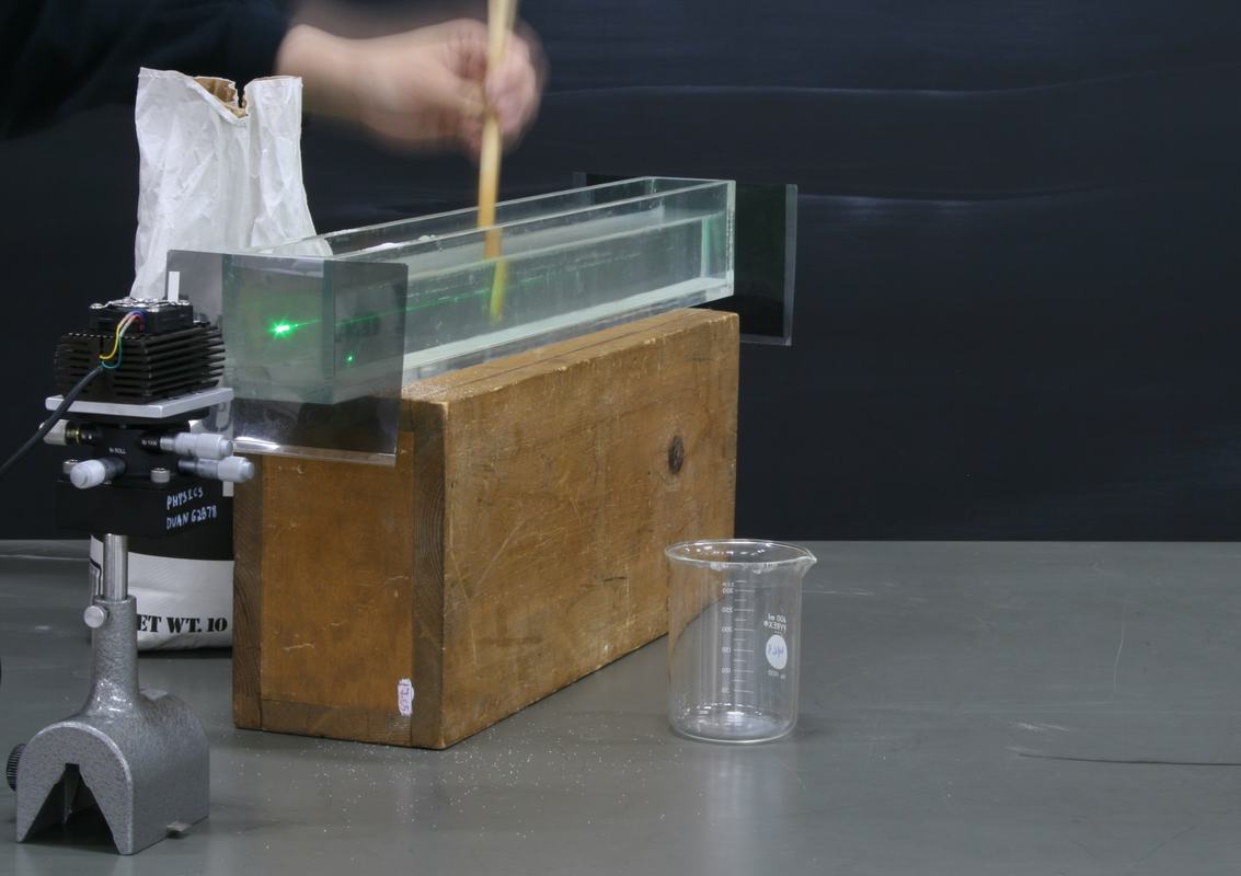 Hand stirring sugar solution in the glass tank with a wooden spoon, laser at left, empty beaker at right
