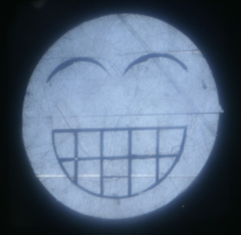 Circular emoji face showing a wide grin with squinted happy eyes, made from Scotch tape strips viewed between crossed polarizers