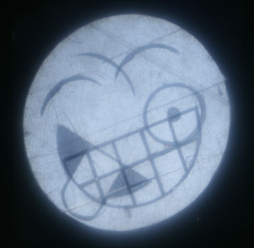 Same emoji face plate rotated 45 degrees, now showing a wild crazy expression with asymmetric eyes and teeth