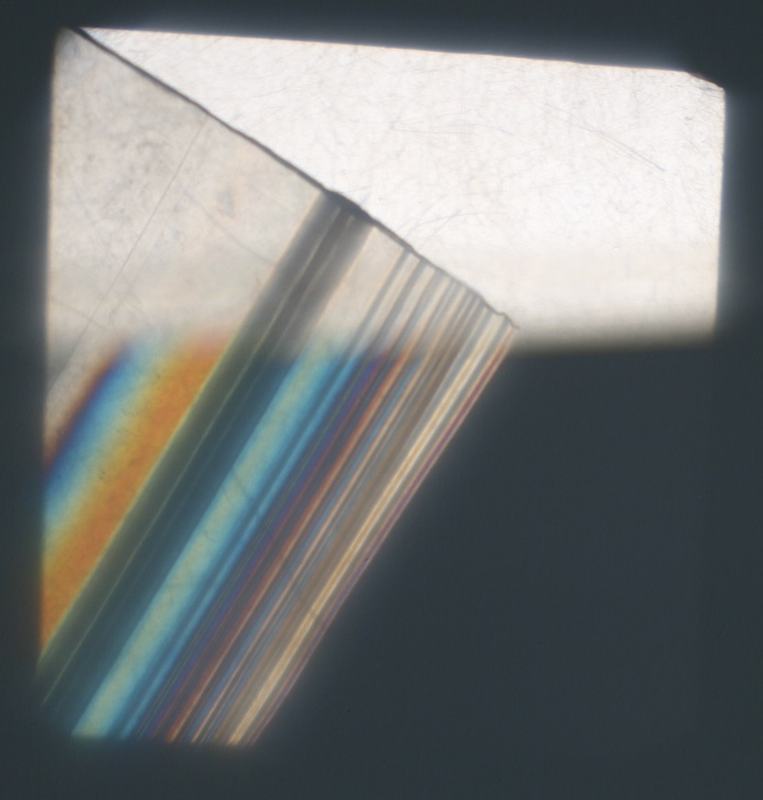 Corner of a zippy bag between crossed polarizers showing parallel diagonal stress fringes in orange, blue, and purple bands