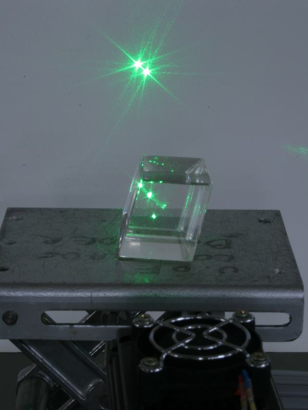 Calcite crystal on a metal platform with green laser beam entering from below, producing two bright green spots on the wall behind
