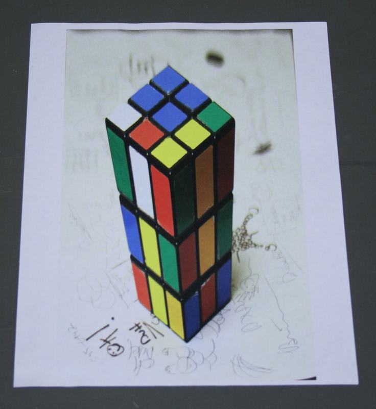Same anamorphic Rubik's cube viewed from above, revealing the elongated distorted shape of the printed image on paper