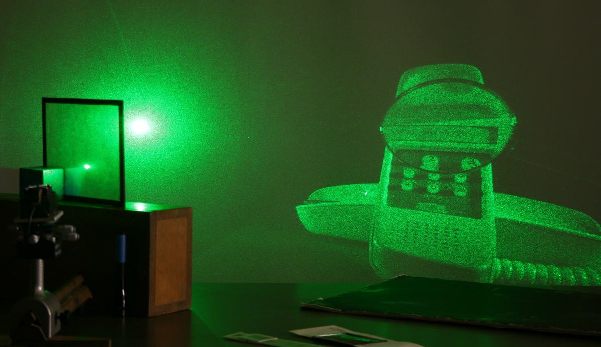 Green laser on wooden stand illuminating a holographic plate, projecting a large holographic image of a telephone onto the wall in a darkened room