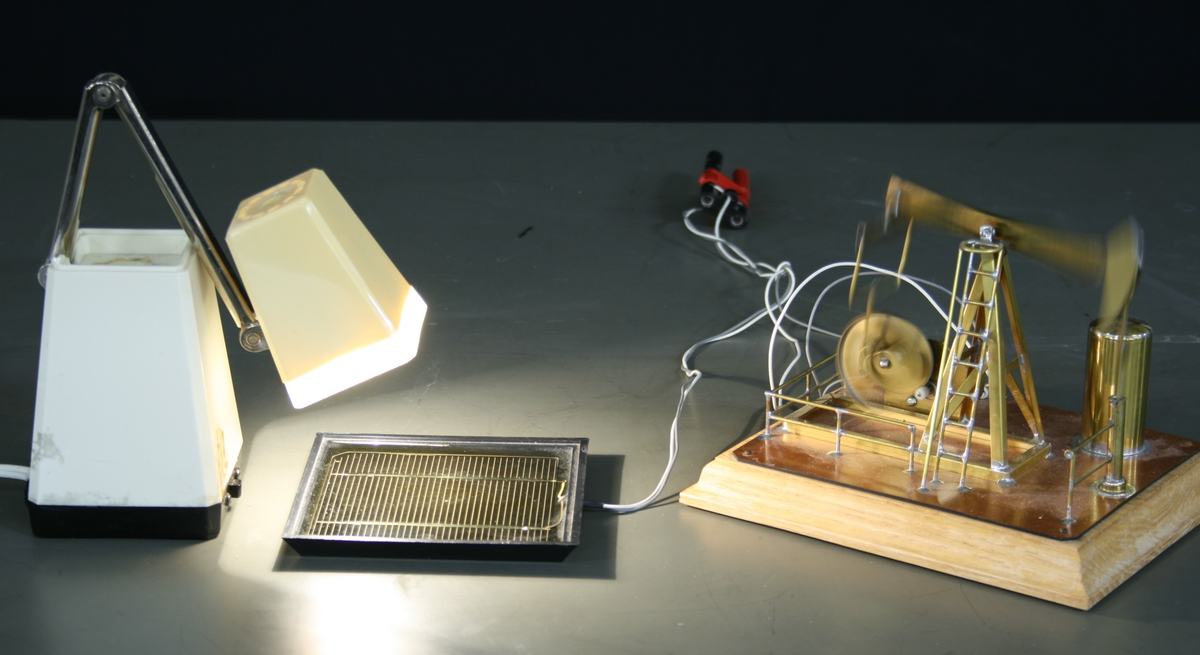 Desk lamp illuminating a solar cell panel connected by wires to a toy oil rig with beam pump mechanism on a wooden base