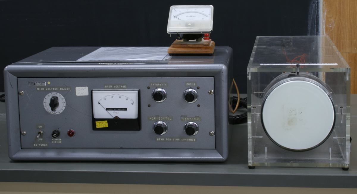 Power supply unit with high voltage adjust dial, analog voltmeter, intensity and focus knobs, and beam position controls, next to evacuated tube in clear acrylic enclosure