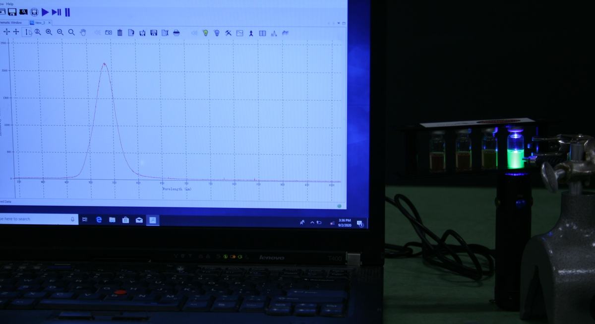 Laptop running OceanView software displaying a fluorescence emission spectrum with a single peak; quantum dot rack with blue LED flashlight and fiber optic cable on ring stand visible at right